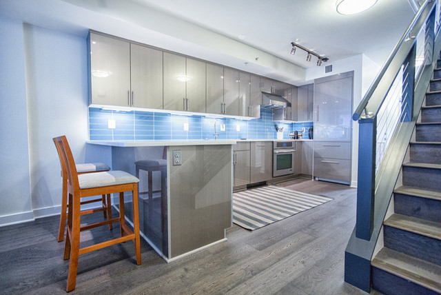 На фото: п-образная кухня-гостиная среднего размера в стиле модернизм с одинарной мойкой, плоскими фасадами, серыми фасадами, столешницей из кварцевого агломерата, синим фартуком, фартуком из стеклянной плитки, техникой из нержавеющей стали, паркетным полом среднего тона и полуостровом