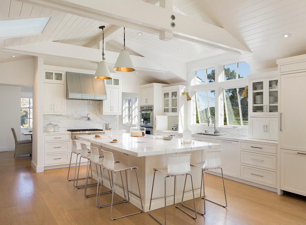 Example of a beach style l-shaped light wood floor kitchen design in San Francisco with an undermount sink, recessed-panel cabinets, white cabinets, marble countertops, white backsplash, stone tile backsplash, paneled appliances and an island