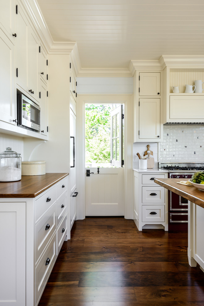 Foto på ett lantligt brun l-kök, med skåp i shakerstil, vita skåp, träbänkskiva, vitt stänkskydd, mellanmörkt trägolv, en köksö och brunt golv