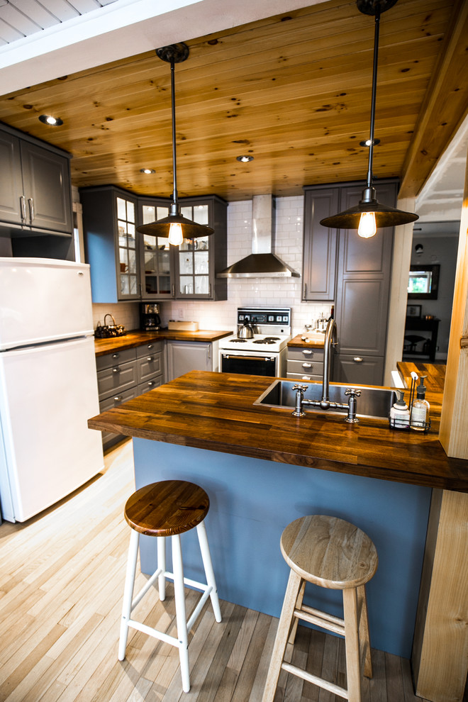 Bild på ett litet rustikt kök, med en enkel diskho, skåp i shakerstil, grå skåp, träbänkskiva, vitt stänkskydd, stänkskydd i porslinskakel, vita vitvaror, ljust trägolv och en köksö