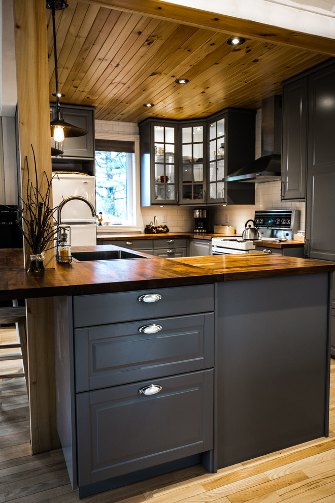 Small rustic l-shaped open plan kitchen in Montreal with a single-bowl sink, shaker cabinets, grey cabinets, wood worktops, white splashback, porcelain splashback, white appliances, light hardwood flooring and an island.