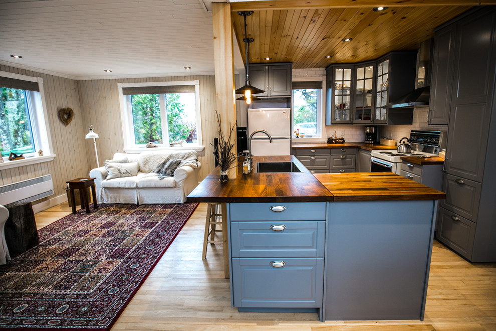 This is an example of a small rustic l-shaped open plan kitchen in Montreal with a single-bowl sink, shaker cabinets, grey cabinets, wood worktops, white splashback, porcelain splashback, white appliances, light hardwood flooring and an island.
