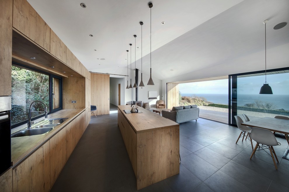 Пример оригинального дизайна: параллельная кухня-гостиная в стиле модернизм с двойной мойкой, плоскими фасадами, фасадами цвета дерева среднего тона, деревянной столешницей, фартуком из стекла, черной техникой и островом