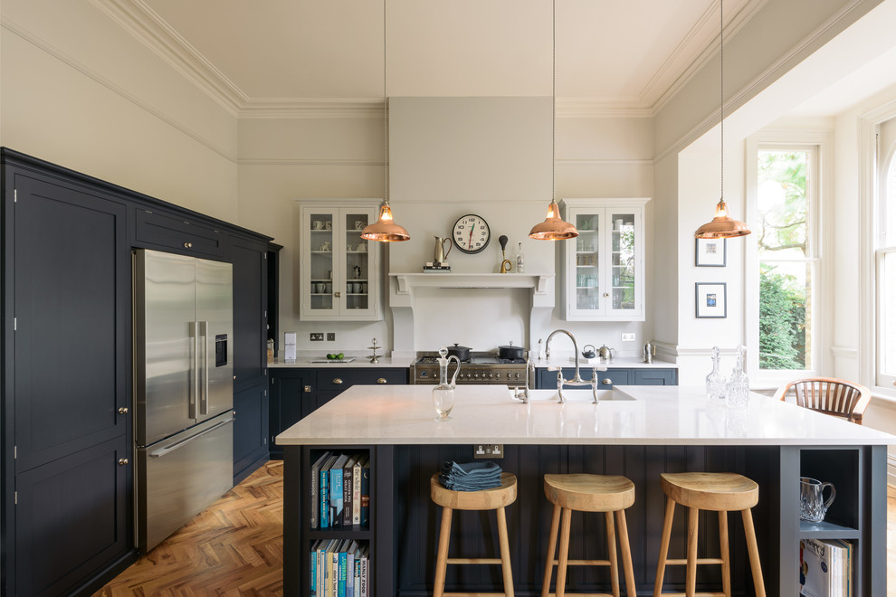 This is an example of a large contemporary l-shaped kitchen/diner in Other with a belfast sink, shaker cabinets, blue cabinets, composite countertops, white splashback, stainless steel appliances, medium hardwood flooring, an island and brown floors.