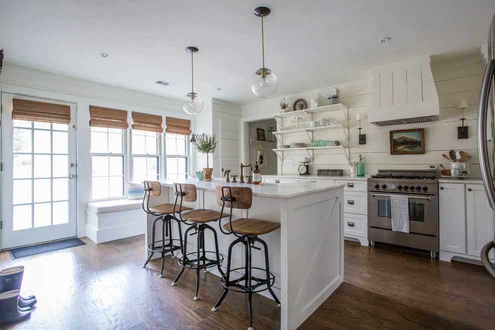 This is an example of a rural l-shaped kitchen in Atlanta with a belfast sink, shaker cabinets, white cabinets, white splashback, stainless steel appliances, dark hardwood flooring and an island.