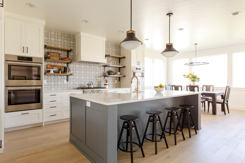 This is an example of a classic galley kitchen/diner in Boise with shaker cabinets, white cabinets, multi-coloured splashback, stainless steel appliances, light hardwood flooring, an island, beige floors and white worktops.