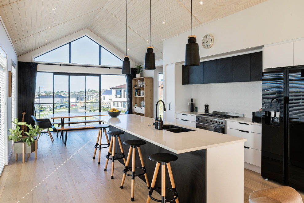 Zweizeilige Moderne Wohnküche mit Doppelwaschbecken, flächenbündigen Schrankfronten, schwarzen Schränken, Küchenrückwand in Weiß, schwarzen Elektrogeräten, hellem Holzboden, Kücheninsel, beigem Boden und weißer Arbeitsplatte in Auckland