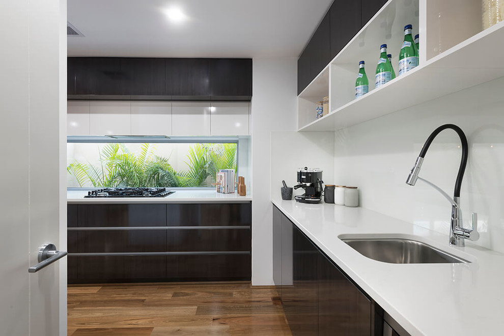 Inspiration for a large contemporary galley open plan kitchen in Perth with a submerged sink, flat-panel cabinets, white cabinets, engineered stone countertops, white splashback, glass sheet splashback, stainless steel appliances, medium hardwood flooring, an island and brown floors.