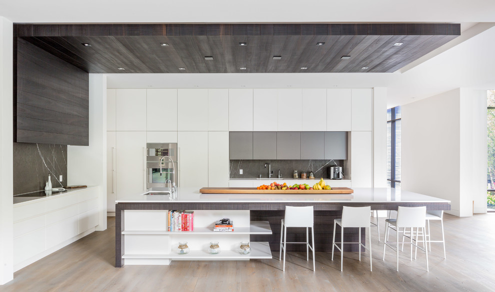 Large contemporary l-shaped open plan kitchen in Toronto with a submerged sink, flat-panel cabinets, white cabinets, engineered stone countertops, grey splashback, stainless steel appliances, light hardwood flooring and an island.