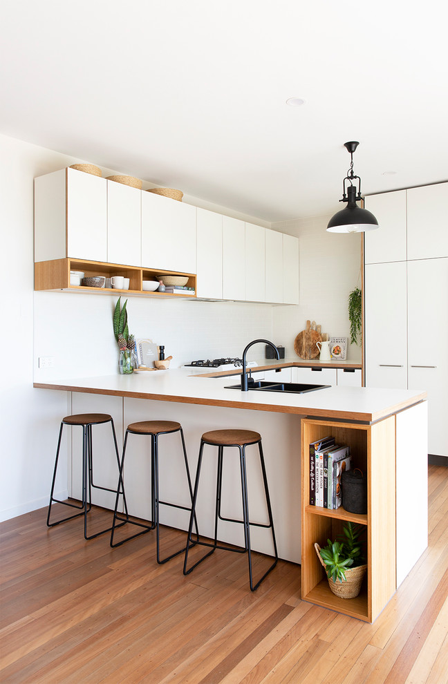 Photo of a nautical u-shaped kitchen in Gold Coast - Tweed with a double-bowl sink, flat-panel cabinets, white cabinets, white splashback, light hardwood flooring, a breakfast bar, beige floors and white worktops.