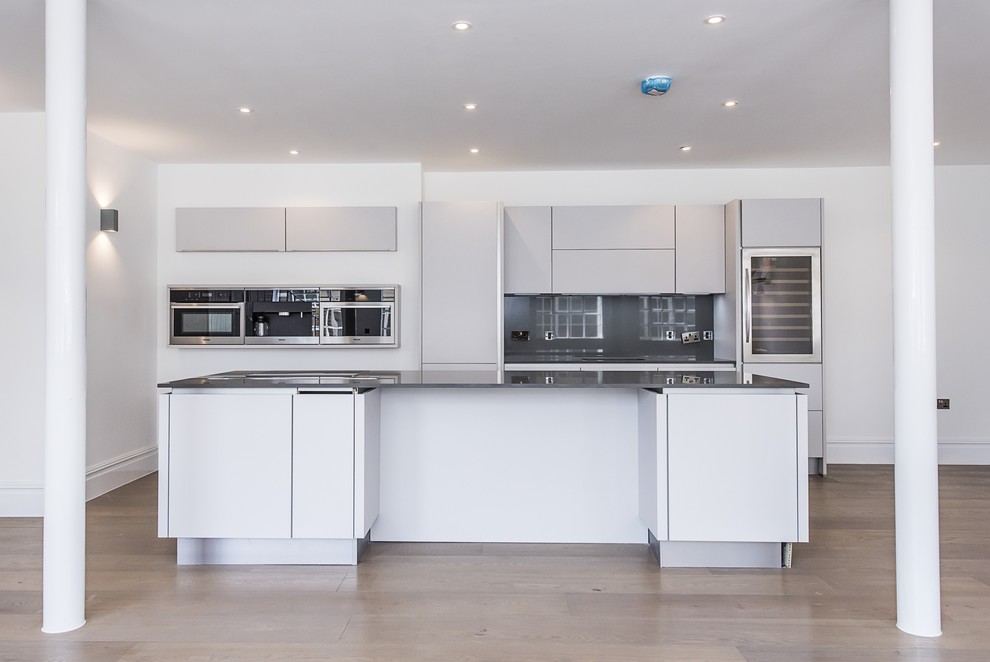 Inspiration for a medium sized contemporary galley open plan kitchen in London with a built-in sink, flat-panel cabinets, white cabinets, grey splashback, stainless steel appliances, medium hardwood flooring and an island.