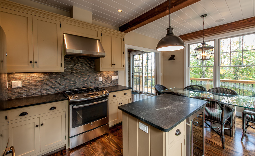 Bild på ett mellanstort lantligt kök, med en undermonterad diskho, luckor med infälld panel, beige skåp, bänkskiva i täljsten, grått stänkskydd, stänkskydd i stenkakel, rostfria vitvaror, mörkt trägolv och en köksö