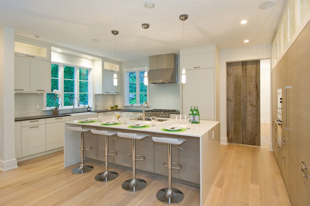 This is an example of a medium sized contemporary l-shaped kitchen/diner in New York with a belfast sink, flat-panel cabinets, white cabinets, white splashback, stainless steel appliances, light hardwood flooring and an island.