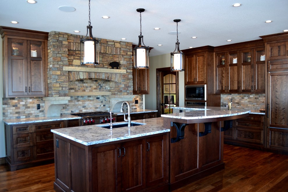 Example of a large tuscan l-shaped dark wood floor and brown floor enclosed kitchen design in Calgary with an undermount sink, recessed-panel cabinets, dark wood cabinets, granite countertops, multicolored backsplash, stone tile backsplash, paneled appliances and an island