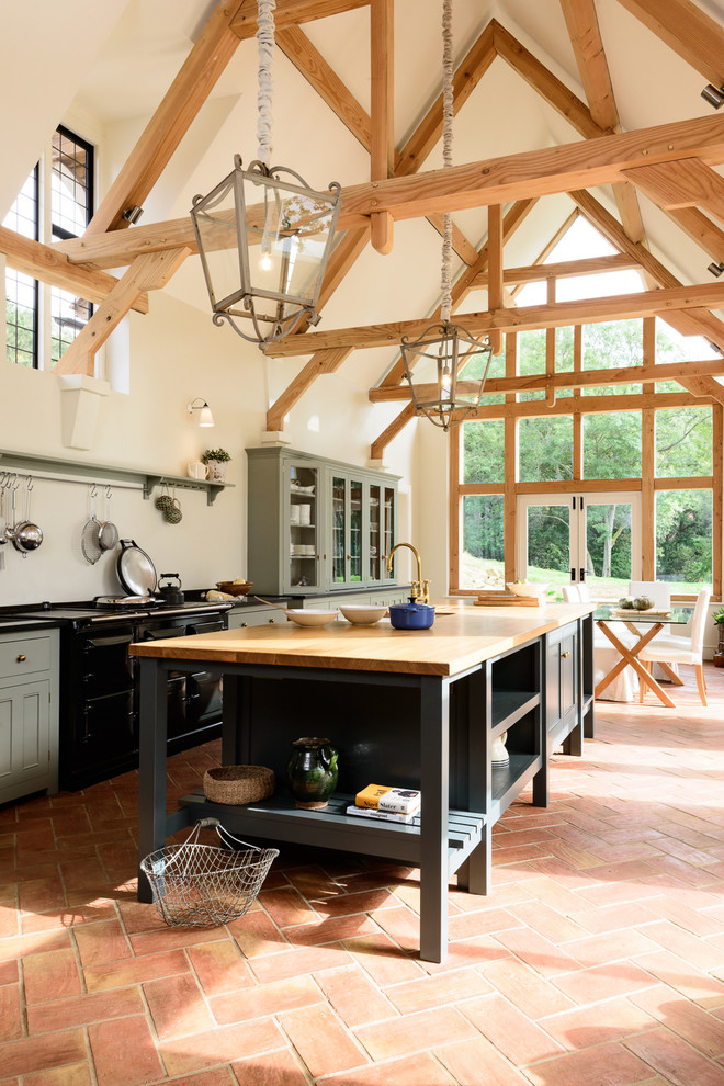 Bild på ett stort lantligt kök och matrum, med grå skåp, svarta vitvaror, klinkergolv i terrakotta, en köksö, luckor med glaspanel och träbänkskiva