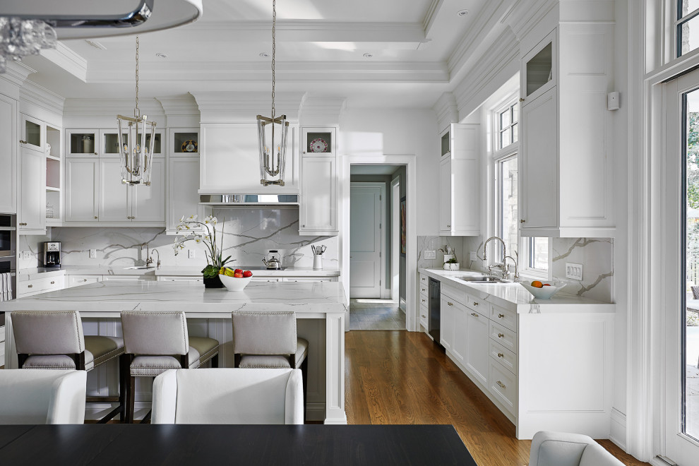 Foto på ett stort vintage vit kök, med en nedsänkt diskho, luckor med infälld panel, vita skåp, marmorbänkskiva, vitt stänkskydd, stänkskydd i marmor, rostfria vitvaror, mellanmörkt trägolv, en köksö och brunt golv