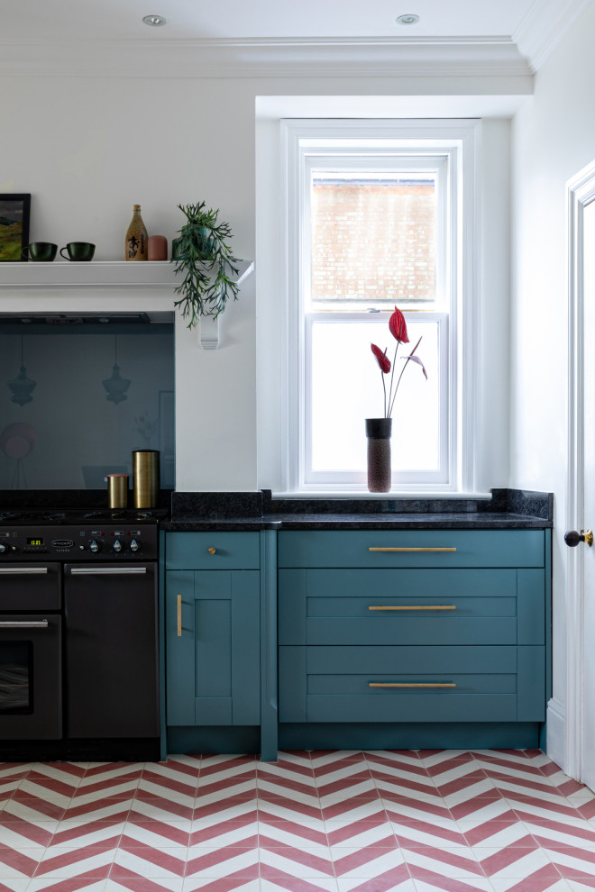 Foto på ett funkis svart kök, med en nedsänkt diskho, skåp i shakerstil, blå skåp, granitbänkskiva, blått stänkskydd, glaspanel som stänkskydd, rostfria vitvaror, cementgolv och rött golv