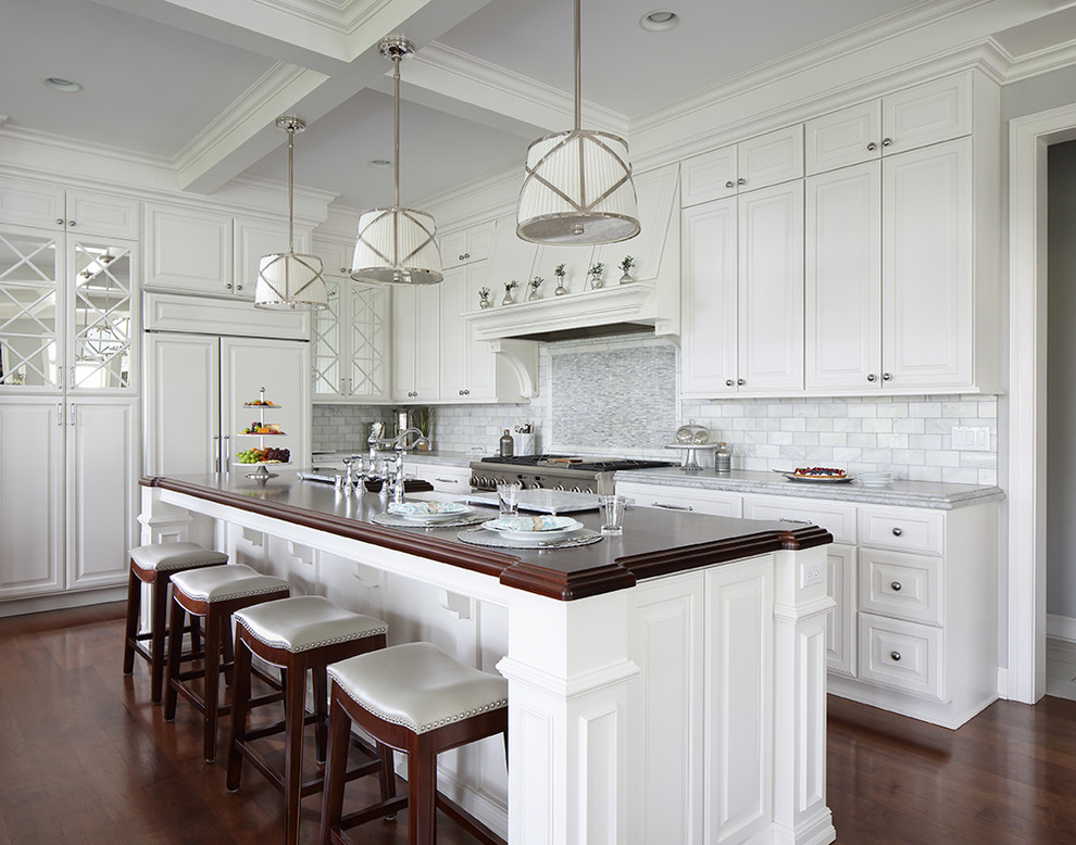 Example of a country l-shaped medium tone wood floor and brown floor kitchen design in Milwaukee with a farmhouse sink, raised-panel cabinets, white cabinets, gray backsplash, paneled appliances, an island and gray countertops