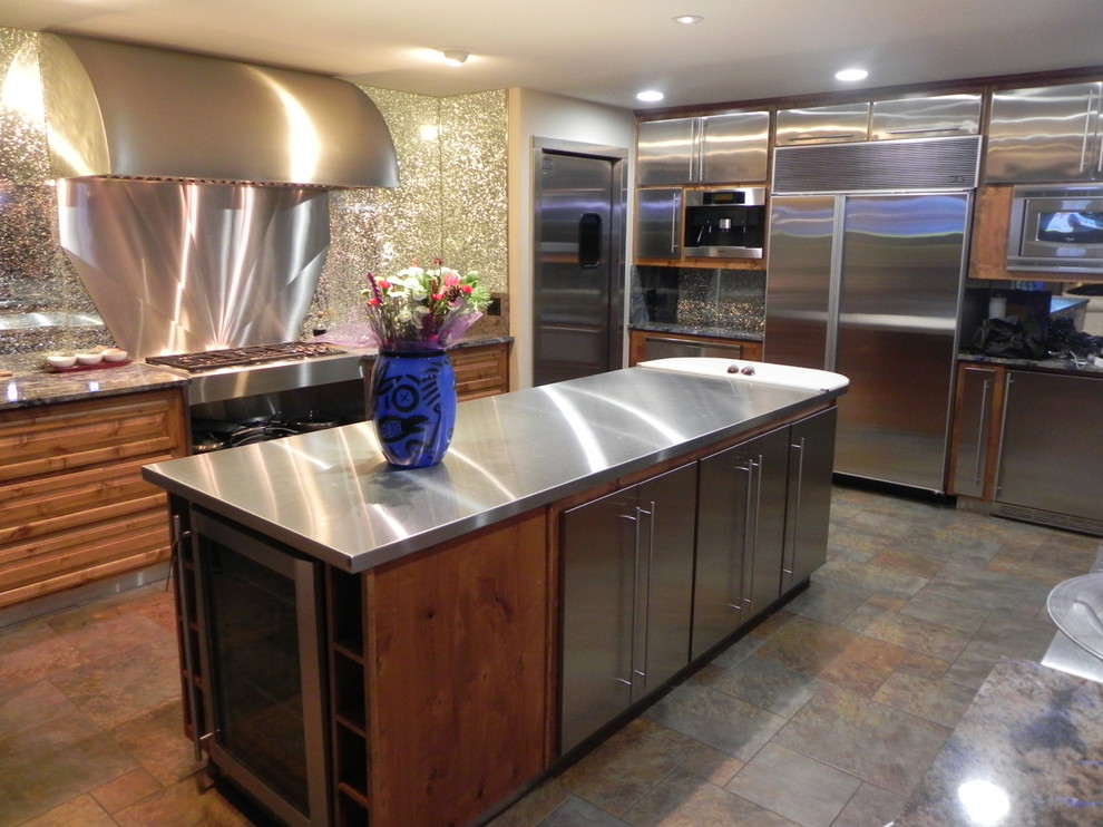 Modern inredning av ett mellanstort l-kök, med släta luckor, skåp i rostfritt stål, bänkskiva i rostfritt stål, glaspanel som stänkskydd, rostfria vitvaror, skiffergolv, en köksö och brunt golv