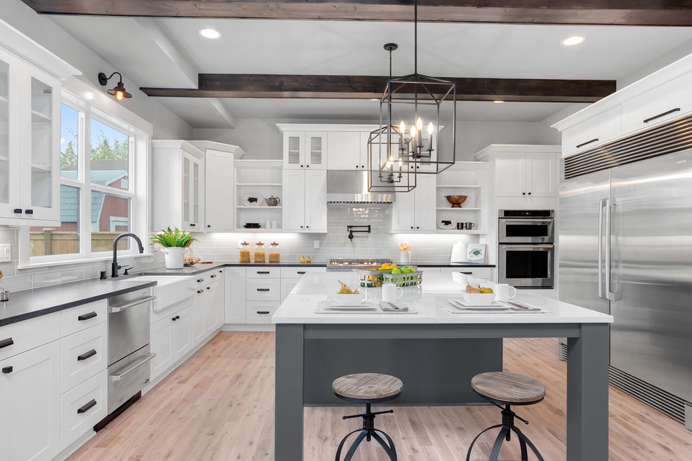 Inspiration for a large farmhouse u-shaped open plan kitchen in Seattle with a belfast sink, shaker cabinets, white cabinets, engineered stone countertops, grey splashback, porcelain splashback, stainless steel appliances, light hardwood flooring, an island and beige floors.