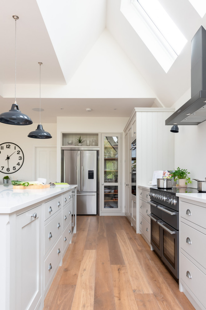 Design ideas for a large classic u-shaped open plan kitchen in Surrey with shaker cabinets, white cabinets, quartz worktops, black appliances, light hardwood flooring, an island, brown floors and white worktops.