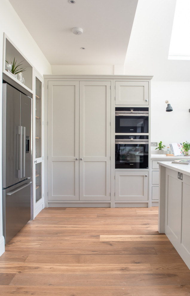 Large traditional u-shaped open plan kitchen in Surrey with shaker cabinets, white cabinets, quartz worktops, black appliances, light hardwood flooring, an island, brown floors and white worktops.
