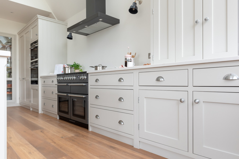 Photo of a large classic u-shaped open plan kitchen in Surrey with shaker cabinets, white cabinets, quartz worktops, black appliances, light hardwood flooring, an island, brown floors and white worktops.