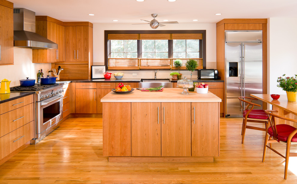 Пример оригинального дизайна: угловая кухня в стиле ретро с техникой из нержавеющей стали, обеденным столом, с полувстраиваемой мойкой (с передним бортиком), плоскими фасадами, фасадами цвета дерева среднего тона и деревянной столешницей