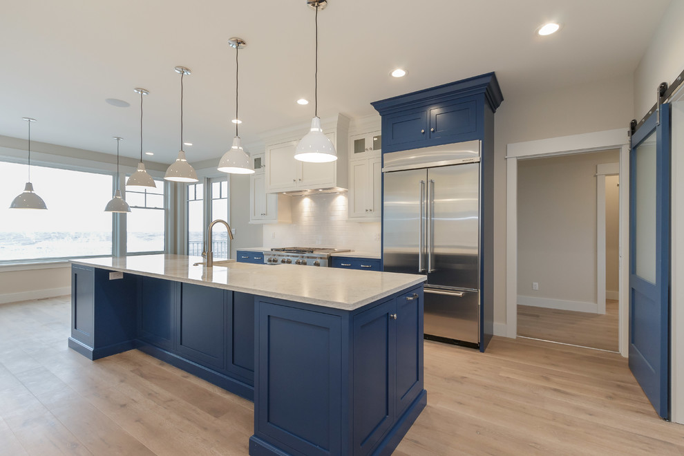 Example of a mid-sized beach style u-shaped light wood floor kitchen pantry design in Other with a farmhouse sink, shaker cabinets, blue cabinets, quartzite countertops, white backsplash, ceramic backsplash, stainless steel appliances and an island