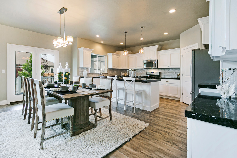 Eat-in kitchen - mid-sized craftsman l-shaped medium tone wood floor and brown floor eat-in kitchen idea in Salt Lake City with a double-bowl sink, recessed-panel cabinets, white cabinets, quartz countertops, white backsplash, porcelain backsplash, stainless steel appliances, an island and black countertops