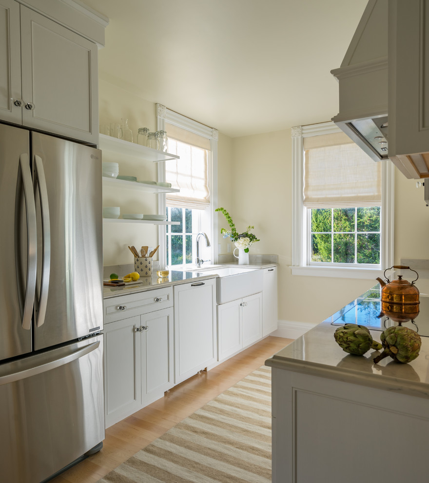 Design ideas for a medium sized farmhouse galley enclosed kitchen in Boston with a belfast sink, shaker cabinets, white cabinets, stainless steel appliances, engineered stone countertops, window splashback, light hardwood flooring, no island and beige floors.