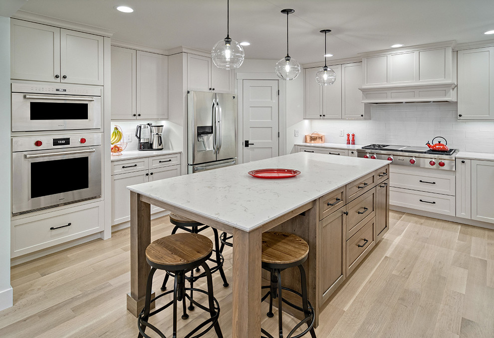 Photo of a large scandinavian l-shaped kitchen/diner in Minneapolis with flat-panel cabinets, white cabinets, engineered stone countertops, white splashback, metro tiled splashback, stainless steel appliances, light hardwood flooring, an island, beige floors, white worktops and a belfast sink.