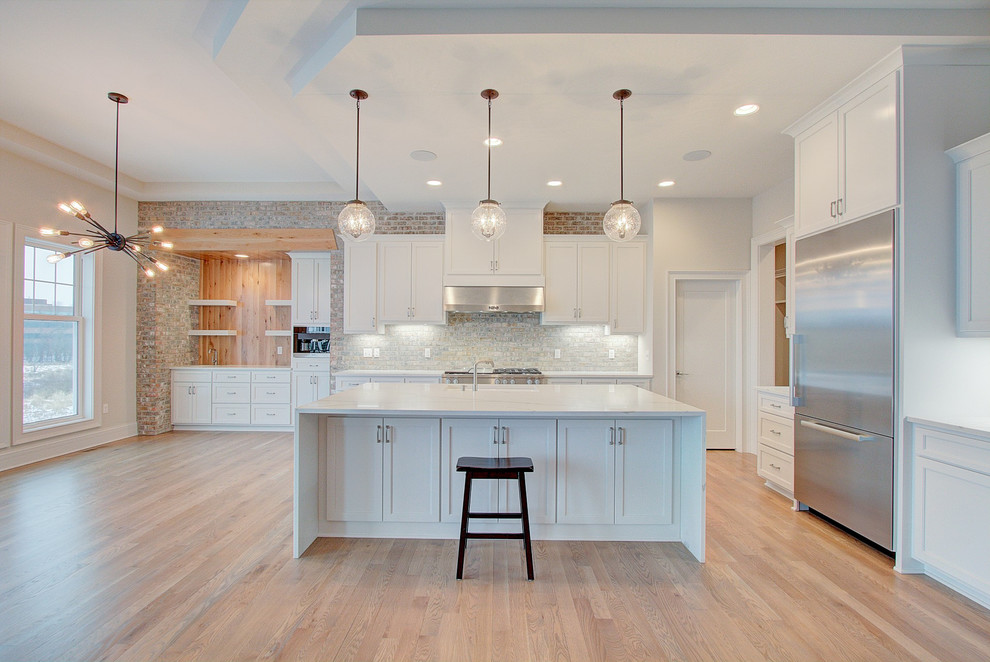Design ideas for a large classic l-shaped open plan kitchen in Milwaukee with shaker cabinets, an island, white cabinets, engineered stone countertops, grey splashback, brick splashback, white worktops, stainless steel appliances, a submerged sink, light hardwood flooring and beige floors.