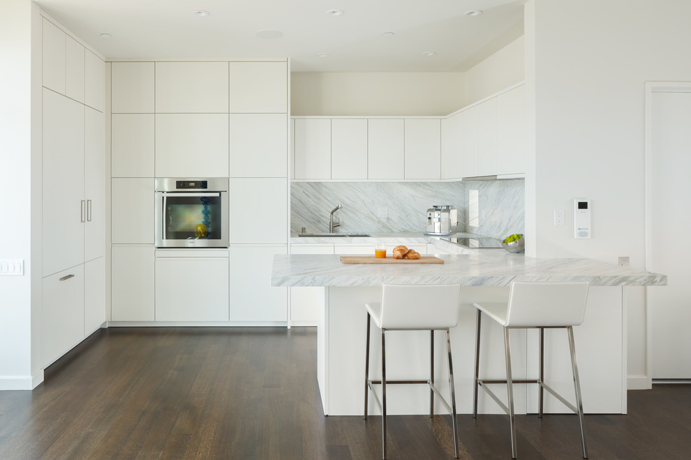 Modern inredning av ett kök med öppen planlösning, med släta luckor, vita skåp, vitt stänkskydd, mörkt trägolv, en halv köksö, en undermonterad diskho, marmorbänkskiva och rostfria vitvaror