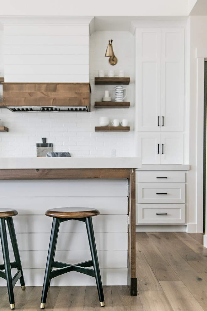 Inspiration for a mid-sized cottage l-shaped medium tone wood floor and brown floor eat-in kitchen remodel in Salt Lake City with a farmhouse sink, shaker cabinets, white cabinets, quartz countertops, white backsplash, subway tile backsplash, stainless steel appliances, an island and white countertops