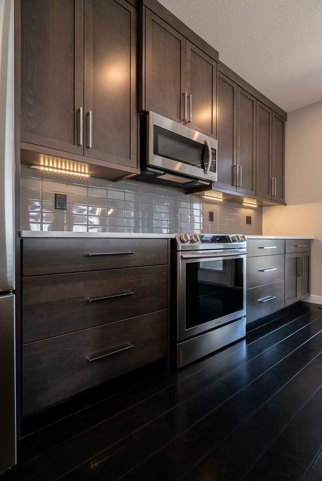 Bild på ett stort amerikanskt vit linjärt vitt kök och matrum, med en undermonterad diskho, skåp i shakerstil, bruna skåp, bänkskiva i kvarts, grått stänkskydd, stänkskydd i tunnelbanekakel, rostfria vitvaror, mörkt trägolv, en köksö och svart golv