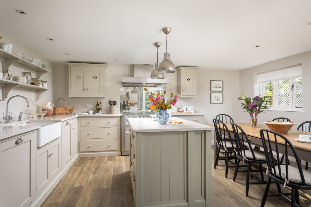 Große Landhausstil Wohnküche in L-Form mit Schrankfronten mit vertiefter Füllung, beigen Schränken, Kücheninsel, Landhausspüle, beiger Arbeitsplatte und hellem Holzboden in Gloucestershire