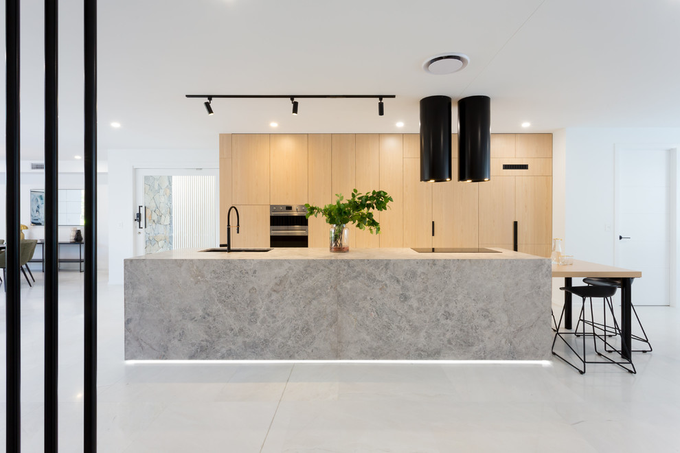 Large contemporary galley open plan kitchen in Brisbane with a submerged sink, light wood cabinets, limestone worktops, black appliances, marble flooring, an island, white floors, grey worktops and flat-panel cabinets.