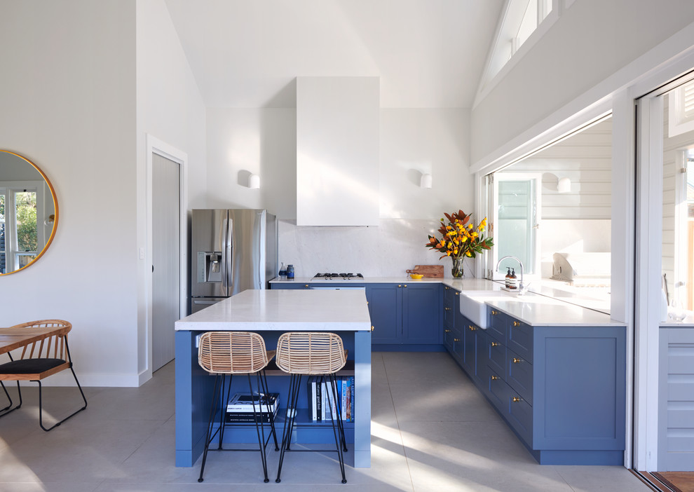 Cette image montre une cuisine traditionnelle en L avec un placard à porte shaker, des portes de placard bleues, îlot, un évier de ferme, une crédence blanche, un électroménager en acier inoxydable et un sol gris.
