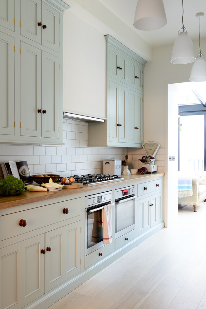 Design ideas for a medium sized traditional galley enclosed kitchen in London with a double-bowl sink, beaded cabinets, blue cabinets, wood worktops, white splashback, metro tiled splashback, stainless steel appliances, light hardwood flooring and no island.