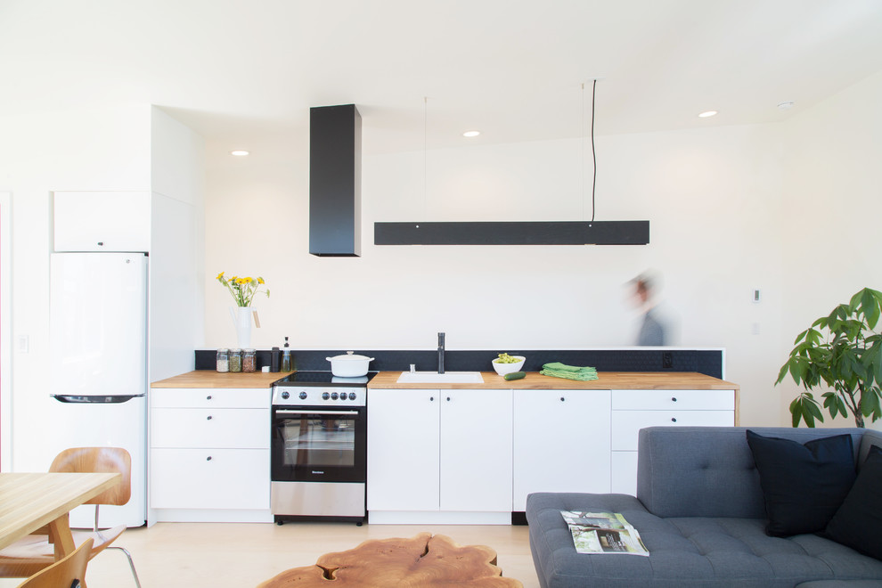 Inspiration för små moderna linjära brunt kök med öppen planlösning, med en enkel diskho, släta luckor, vita skåp, träbänkskiva, svart stänkskydd, vita vitvaror, plywoodgolv och vitt golv