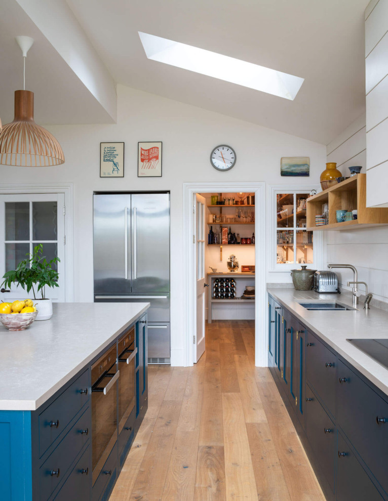 Inredning av ett modernt mellanstort vit vitt kök med öppen planlösning, med en nedsänkt diskho, skåp i shakerstil, blå skåp, bänkskiva i kvartsit, ljust trägolv och en köksö