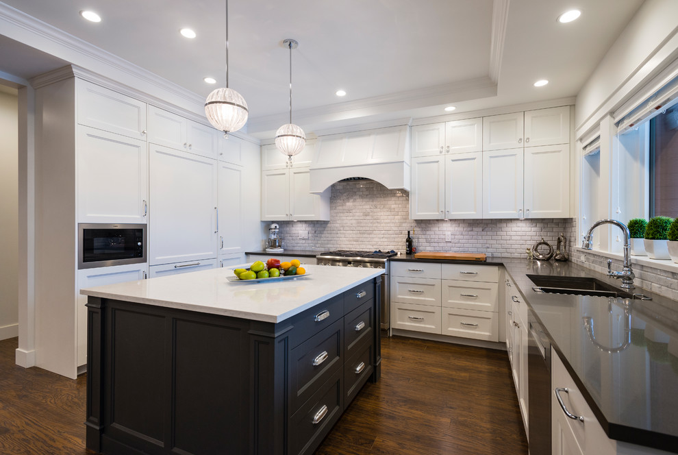 Klassisk inredning av ett mellanstort kök, med en undermonterad diskho, luckor med infälld panel, vita skåp, bänkskiva i kvartsit, grått stänkskydd, stänkskydd i stenkakel, rostfria vitvaror, mörkt trägolv, en köksö och brunt golv