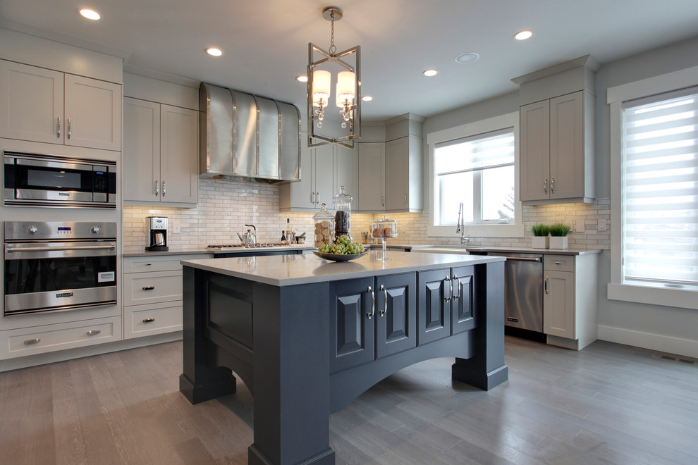 Inspiration for a transitional l-shaped kitchen remodel in Calgary with a farmhouse sink, shaker cabinets, gray cabinets, white backsplash, stainless steel appliances, an island and stone tile backsplash