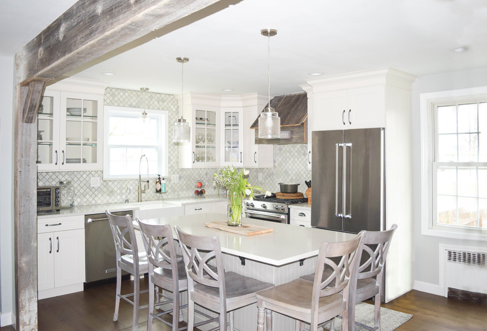 This is an example of a medium sized shabby-chic style l-shaped kitchen/diner in New York with a belfast sink, glass-front cabinets, white cabinets, engineered stone countertops, grey splashback, stone tiled splashback, stainless steel appliances, dark hardwood flooring and an island.