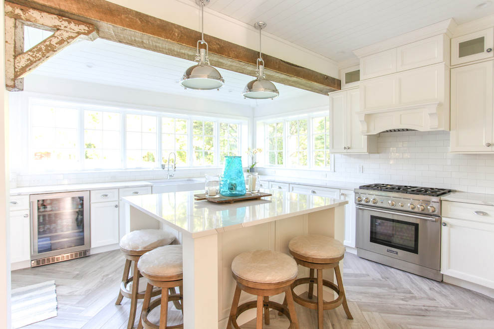 This is an example of a large beach style u-shaped kitchen/diner in New York with a belfast sink, shaker cabinets, white cabinets, engineered stone countertops, white splashback, metro tiled splashback, integrated appliances, porcelain flooring, an island, grey floors and white worktops.