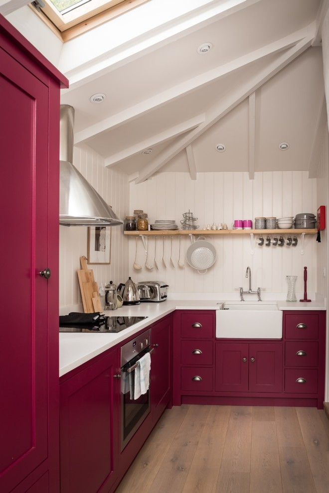 This is an example of a small rural l-shaped enclosed kitchen in Cornwall with a belfast sink, shaker cabinets, red cabinets, laminate countertops, white splashback, wood splashback, stainless steel appliances, medium hardwood flooring and beige floors.