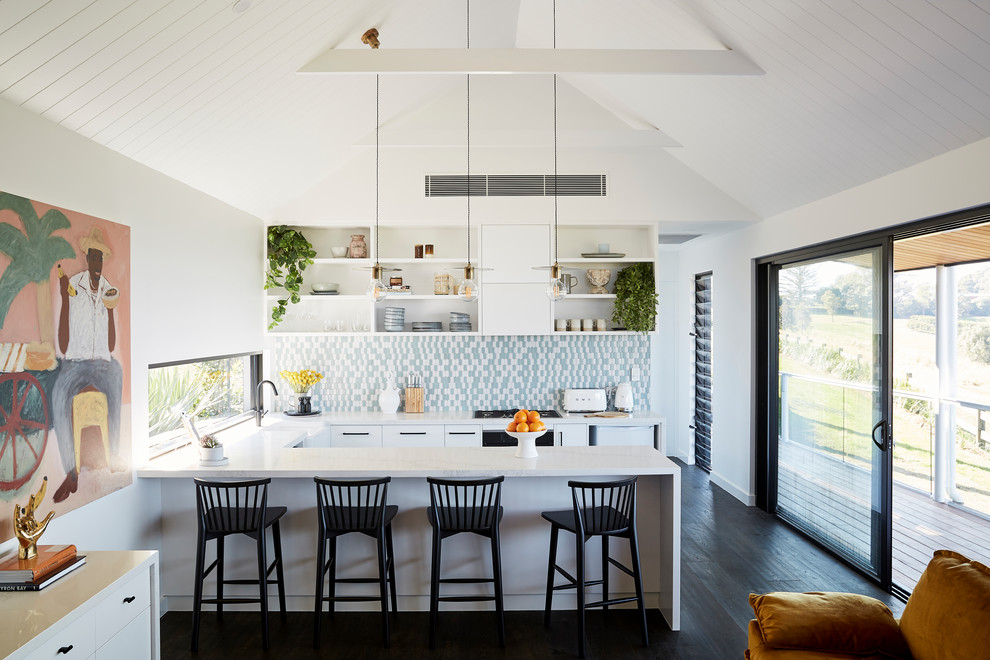 Coastal u-shaped open plan kitchen in Other with a belfast sink, flat-panel cabinets, white cabinets, multi-coloured splashback, integrated appliances, dark hardwood flooring, a breakfast bar, black floors and white worktops.