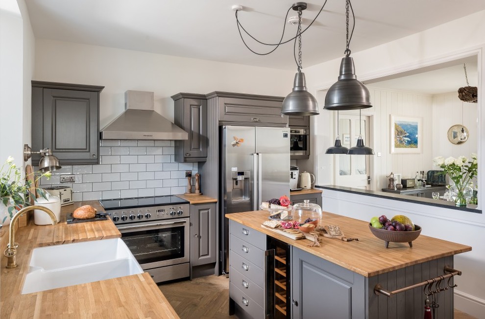 Inspiration for a nautical l-shaped kitchen/diner in Cornwall with a double-bowl sink, raised-panel cabinets, grey cabinets, wood worktops, white splashback, metro tiled splashback, stainless steel appliances, medium hardwood flooring, an island, brown floors and brown worktops.