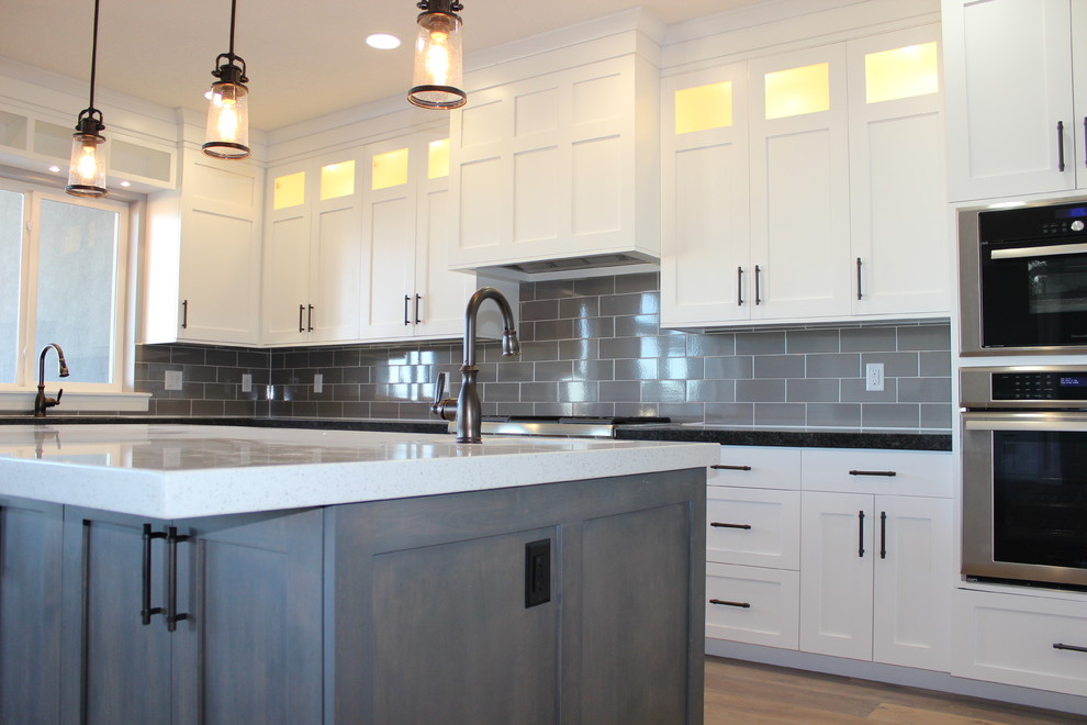 Foto på ett stort amerikanskt kök, med en undermonterad diskho, luckor med upphöjd panel, vita skåp, granitbänkskiva, grått stänkskydd, stänkskydd i keramik, rostfria vitvaror, mellanmörkt trägolv och en köksö